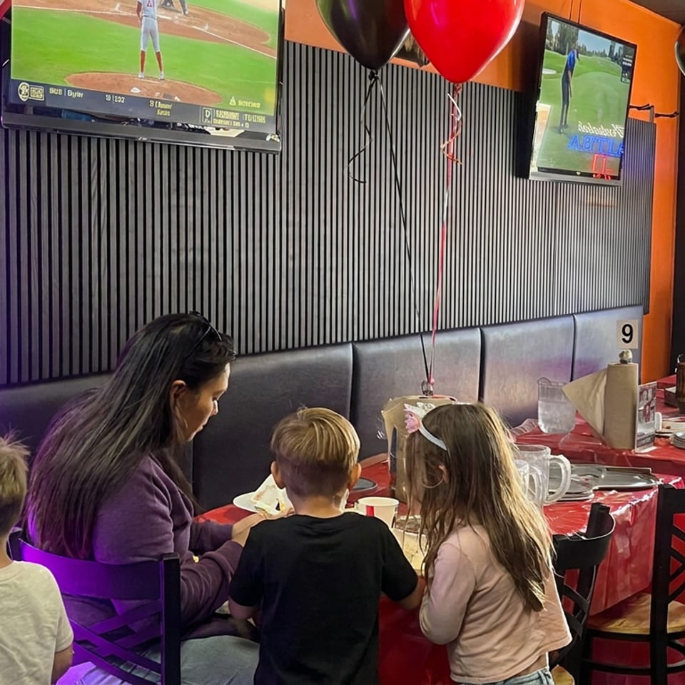 Family gathers at a decorated restaurant table beside a prize machine, with sports playing on TVs and red, black, and white balloons.