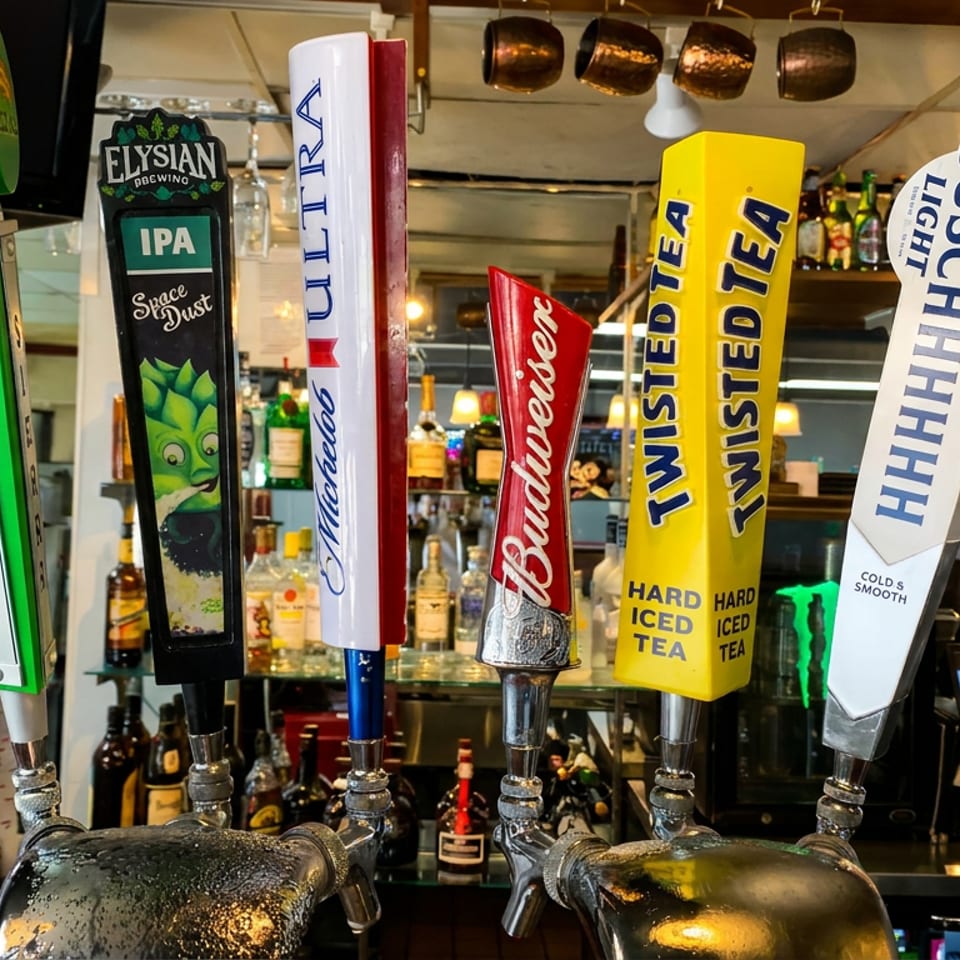 Colorful beer and hard iced tea tap handles line a busy bar, with liquor bottles, glassware, and a cash register behind them.