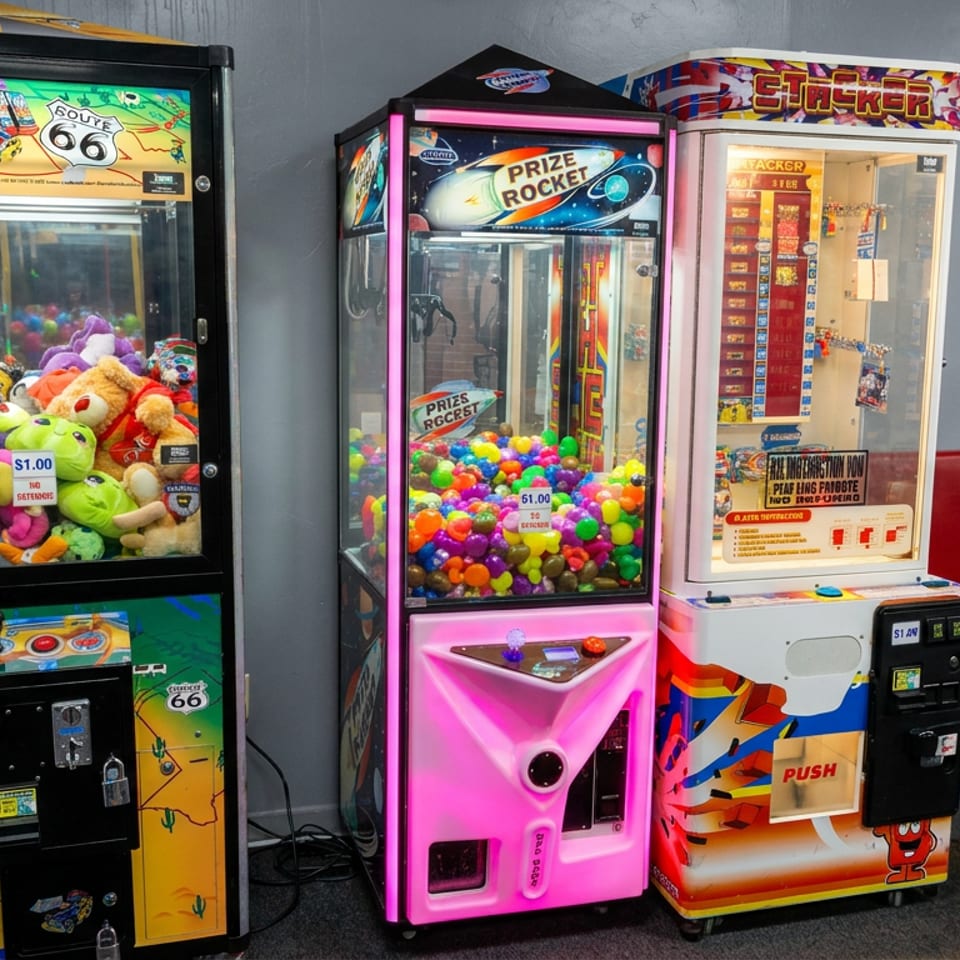 Three colorful arcade prize machines filled with plush toys and plastic balls stand beside red restaurant booths and tables.