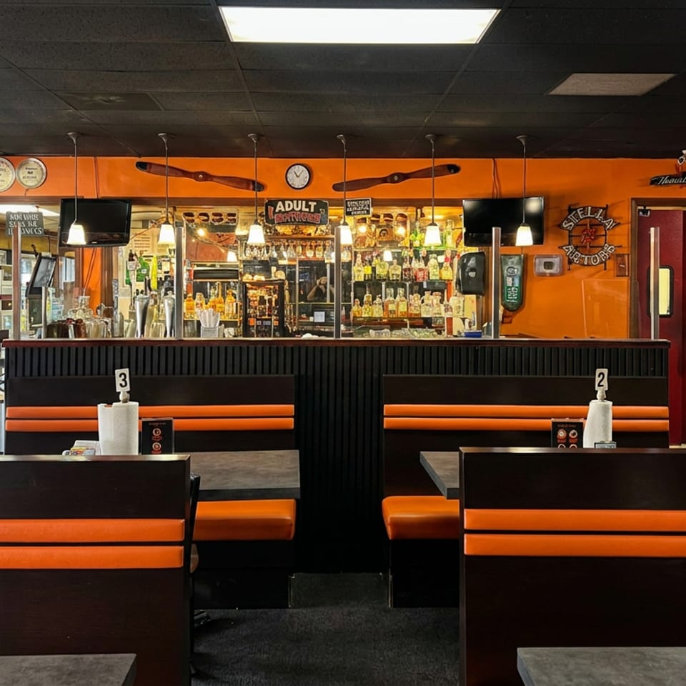 Retro restaurant interior with orange-and-black booths, a stocked bar, pendant lights, and vintage decor.